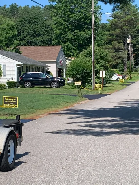 A residential street with houses, parked cars, and trees. There are signs on the lawns, including one advertising Young's Hot Top Paving with a phone number.