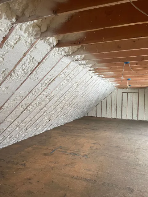 Attic with angled ceiling and walls insulated with white spray foam. Wooden beams and unfinished floor.