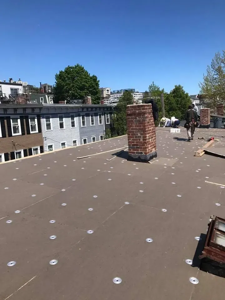 Rooftop with a brick chimney, fasteners, and a worker in the distance, surrounded by buildings on a sunny day.