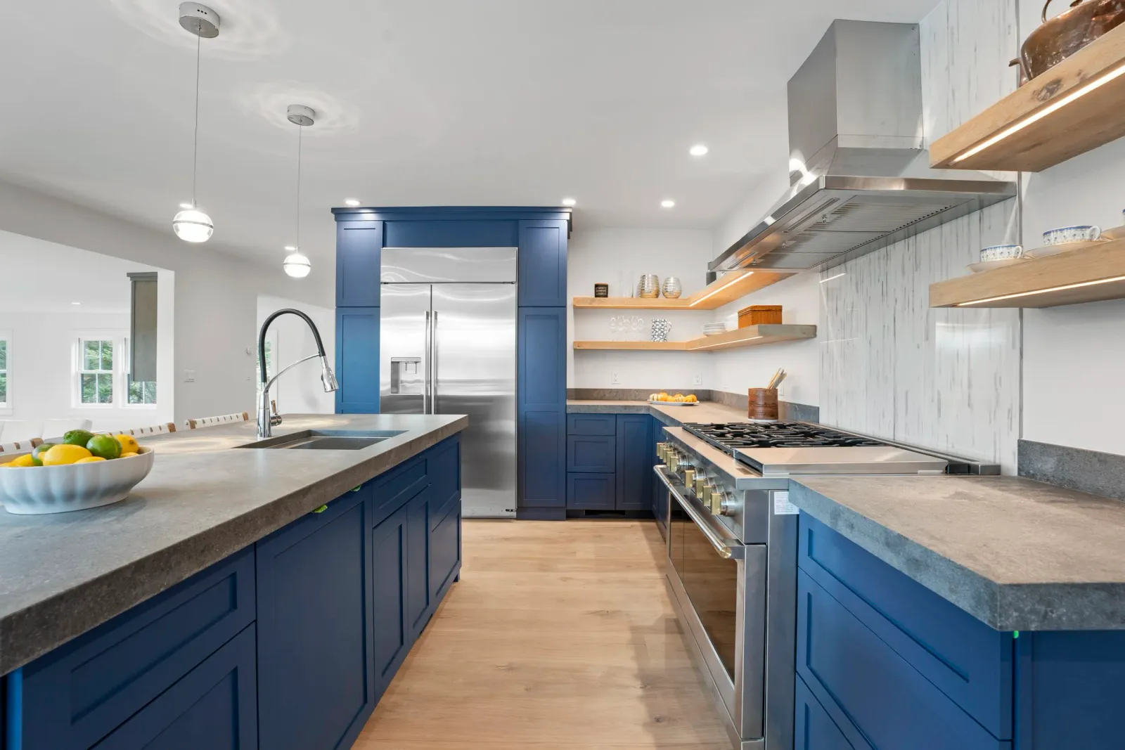Modern kitchen with blue cabinets, stainless steel appliances, wooden open shelves, and a central island with a bowl of lemons and limes.