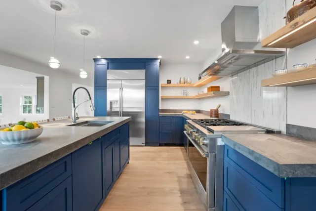 Modern kitchen with blue cabinets, stainless steel appliances, wooden open shelves, and a central island with a bowl of lemons and limes.
