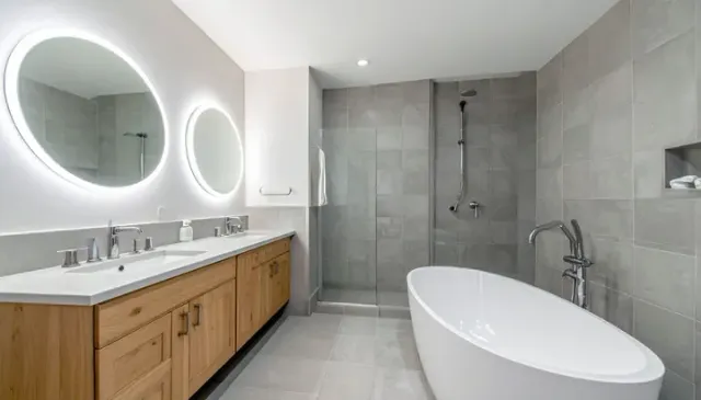 A modern bathroom with a light wood double vanity, two round lighted mirrors, a white oval tub, and a glass-walled shower.