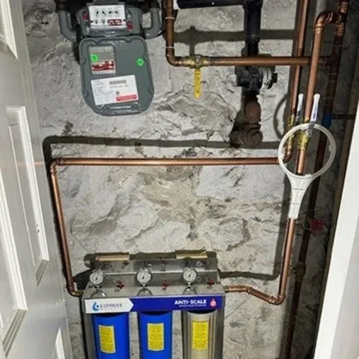 Copper pipes and a water filtration system with three blue filters are mounted on a stone basement wall. An anti-scale label is visible, along with a large white filter wrench hanging on a pipe.
