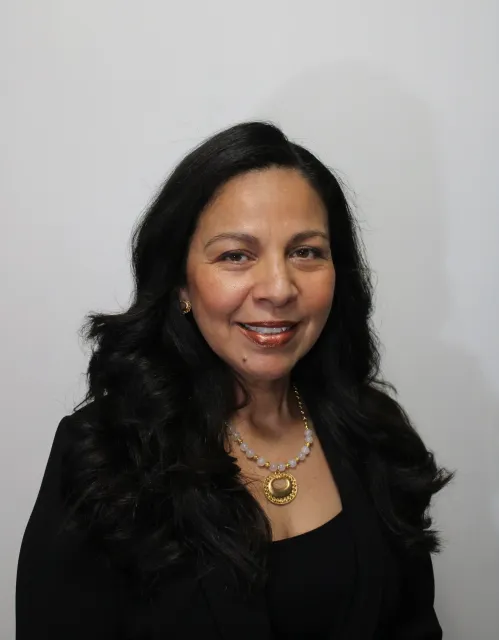Portrait of a woman with long black hair wearing a black blazer, pearl necklace with a gold pendant, and gold earrings, smiling in front of a plain white wall.
