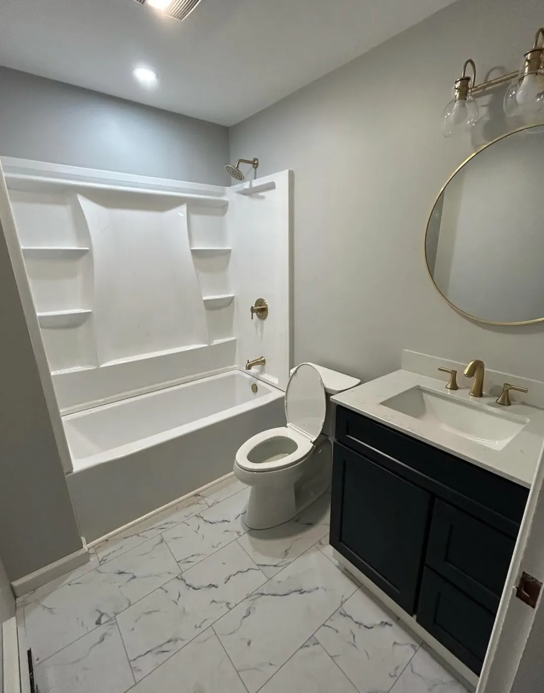 Modern bathroom with a white bathtub and shower, a toilet, sink with a dark vanity, round mirror, and light fixture, marble-patterned flooring.