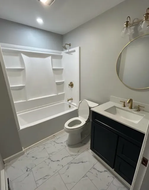 Modern bathroom with a white bathtub and shower, a toilet, sink with a dark vanity, round mirror, and light fixture, marble-patterned flooring.