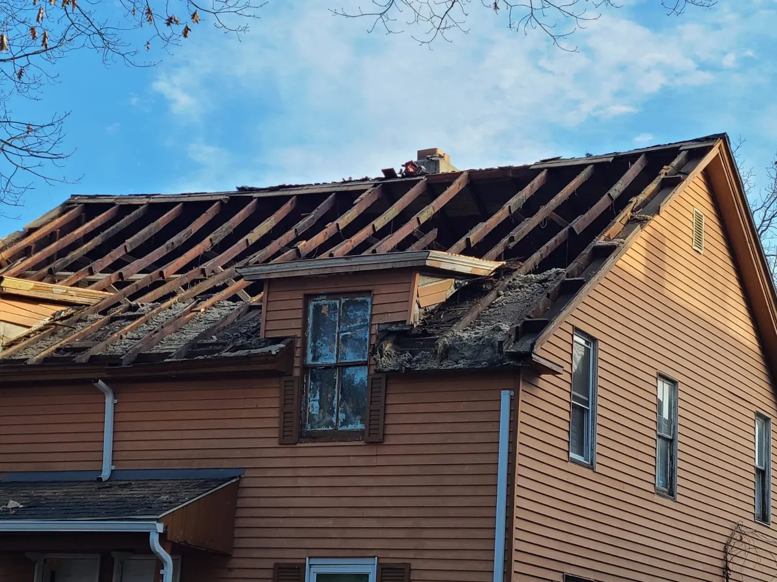 The roof of a house with significant damage and missing shingles, revealing the underlying structure and insulation. The house has beige siding and multiple windows, with a partly cloudy sky in the background.