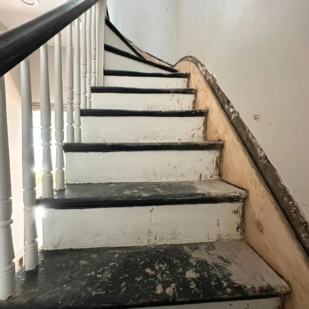 A weathered staircase with black painted treads and white risers, with a black handrail on the left side, in a partially renovated or old building.