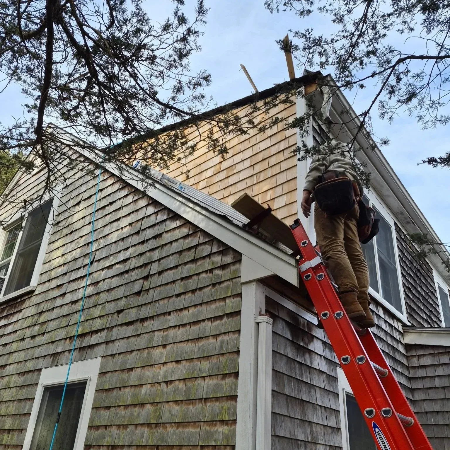 A house with shingle siding under repair, a worker standing on a red ladder is fixing the roof, with trees and blue sky in the background.