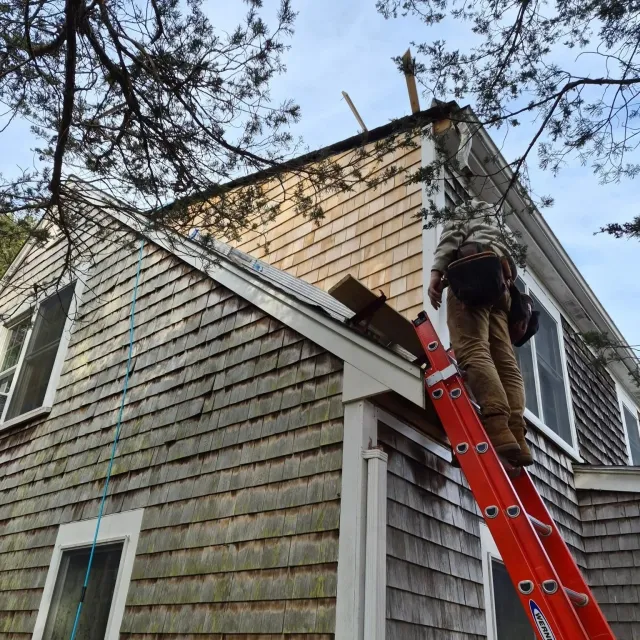A house with shingle siding under repair, a worker standing on a red ladder is fixing the roof, with trees and blue sky in the background.