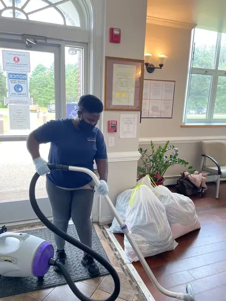 Person vacuuming a lobby floor beside bags and a potted plant near a window.