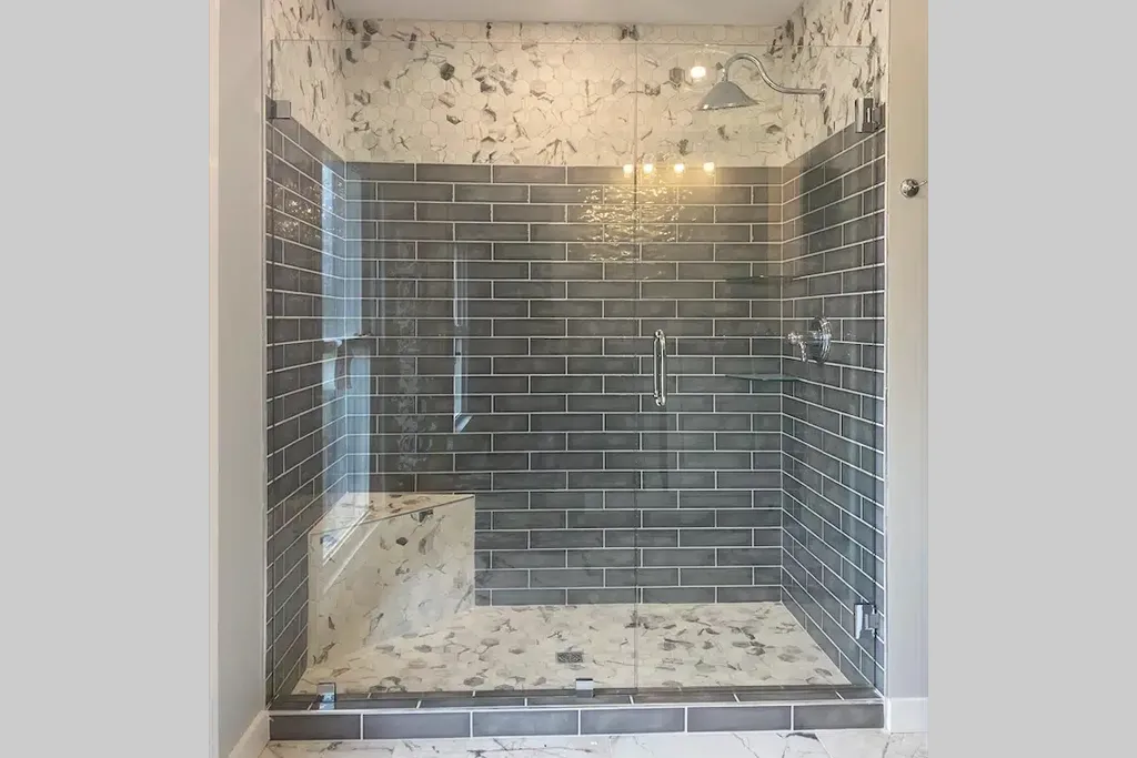 A modern walk-in shower with dark gray subway tiles on the walls, white stone tiles with gray accents on the ceiling and floor, a glass door, a built-in seat in the corner, and a showerhead and controls mounted on the wall.