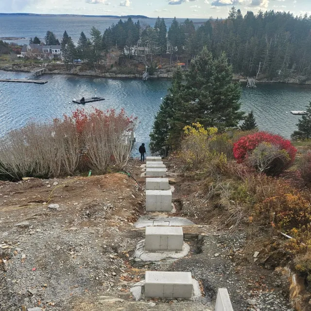 Construction site on a hillside overlooking a river with trees, houses, and boats in the background, and a person walking down the path.