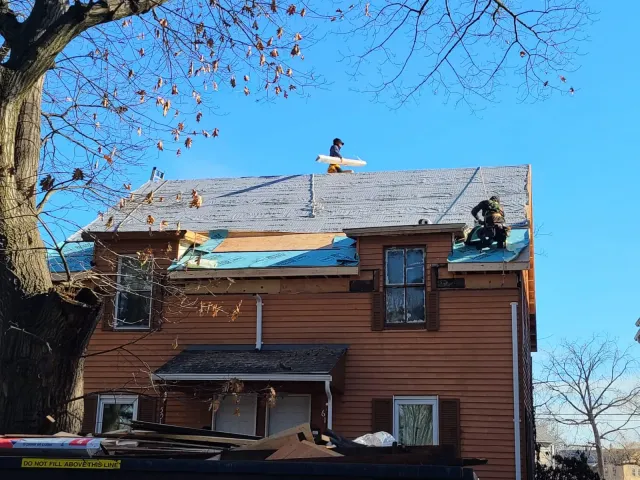 Two construction workers on the roof of a house, installing or repairing shingles, with a clear blue sky and leafless trees in the background.