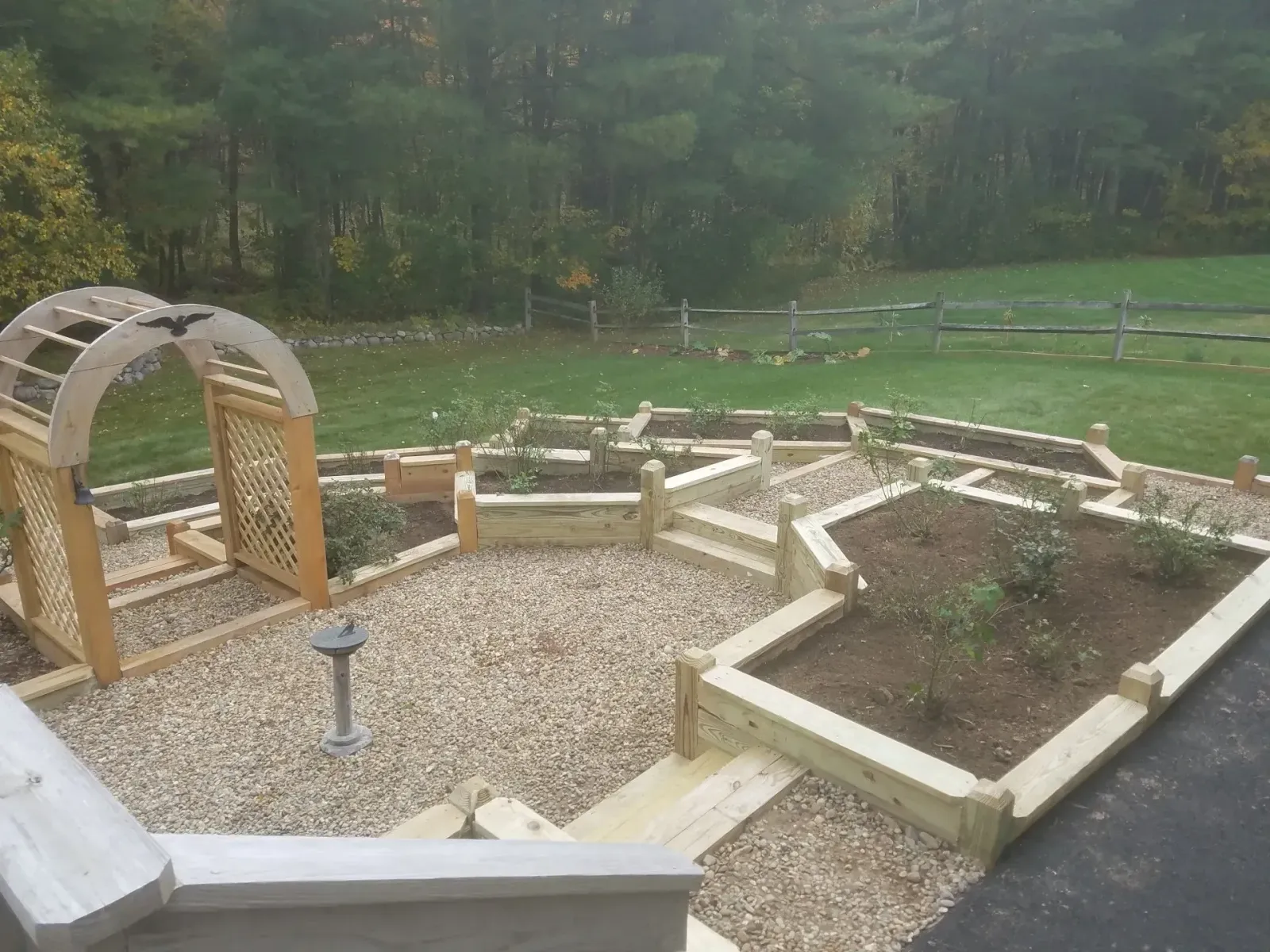 Raised garden beds and gravel paths beside a wooden arched trellis in a landscaped backyard.