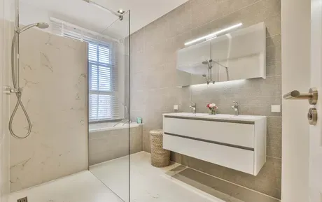 Modern beige bathroom with glass shower, white vanity, mirror, and window blinds