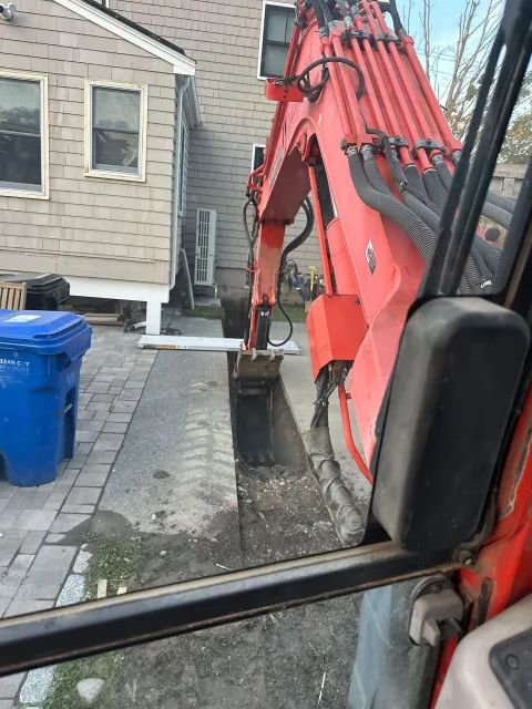 A view from the cab of an orange excavator digging a narrow trench alongside a house next to a blue trash bin.