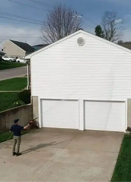 A man performs exterior cleaning on a white two-car garage with a power washer, showcasing home maintenance.