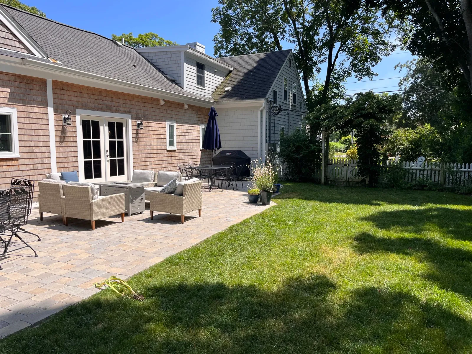 A backyard patio with wicker and metal furniture, a grill, potted plants, and a white picket fence surrounded by green grass and trees, under a blue sky.