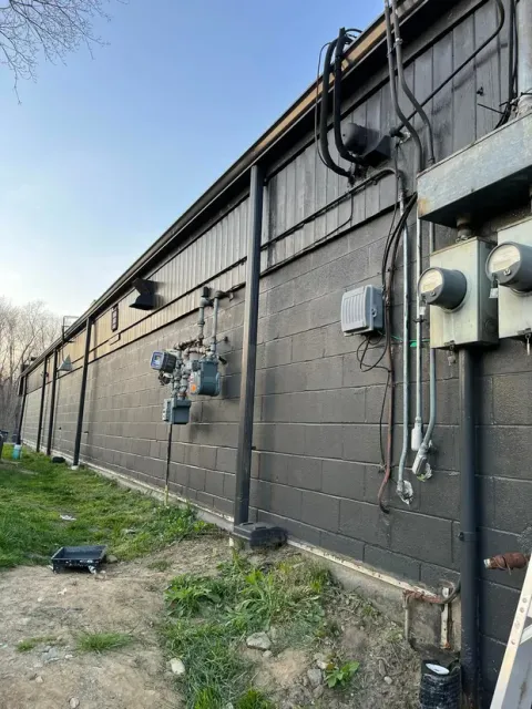 Black building exterior with electrical boxes, pipes, and meters. Green grass in front, blue sky.