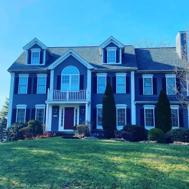 A large, three-story colonial-style house painted in navy blue with white trim and black shutters. The house has a prominent central arched window and dormer windows on the roof. The front yard has a well-maintained lawn with tall, narrow bushes and