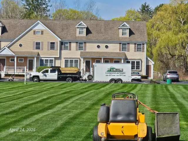 Lawn care crew working outside a beige house, with trucks and a yellow mower on striped grass.