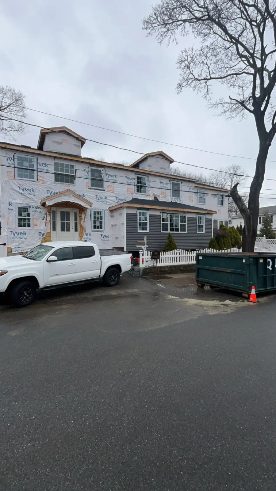 A house under construction with Tyvek sheathing on the exterior, a white pickup truck parked in front, and a large green dumpster on the driveway, with a tree and overcast sky in the background.