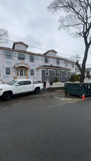 A house under construction with Tyvek sheathing on the exterior, a white pickup truck parked in front, and a large green dumpster on the driveway, with a tree and overcast sky in the background.