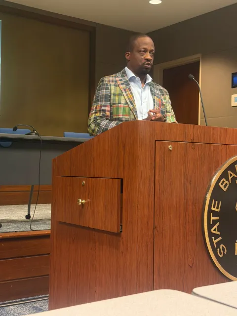 Man speaking at a wooden podium in a courtroom or hearing room, wearing a plaid jacket.