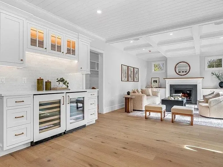 Hardwood floor installation in a Plymouth living room with white walls, a fireplace, light-colored furniture, and an adjacent kitchen with white cabinets and a wine fridge.