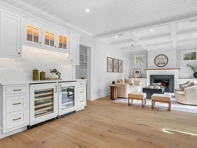 Hardwood floor installation in a Plymouth living room with white walls, a fireplace, light-colored furniture, and an adjacent kitchen with white cabinets and a wine fridge.