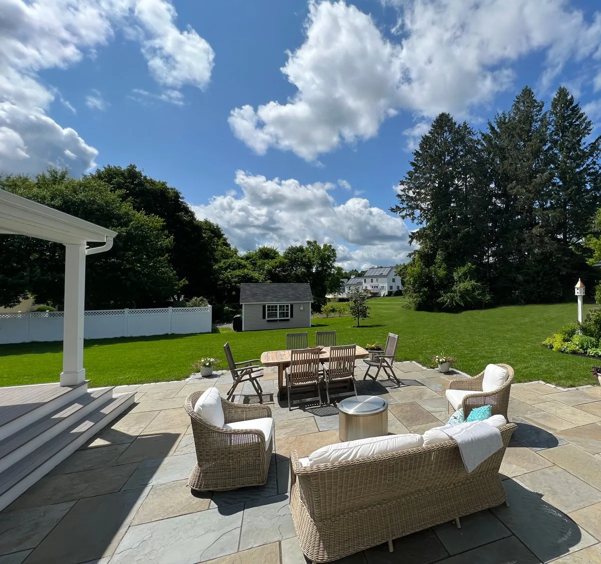 Spacious patio with wicker furniture overlooks lush backyard, trees, and a small shed under a blue sky with scattered clouds.