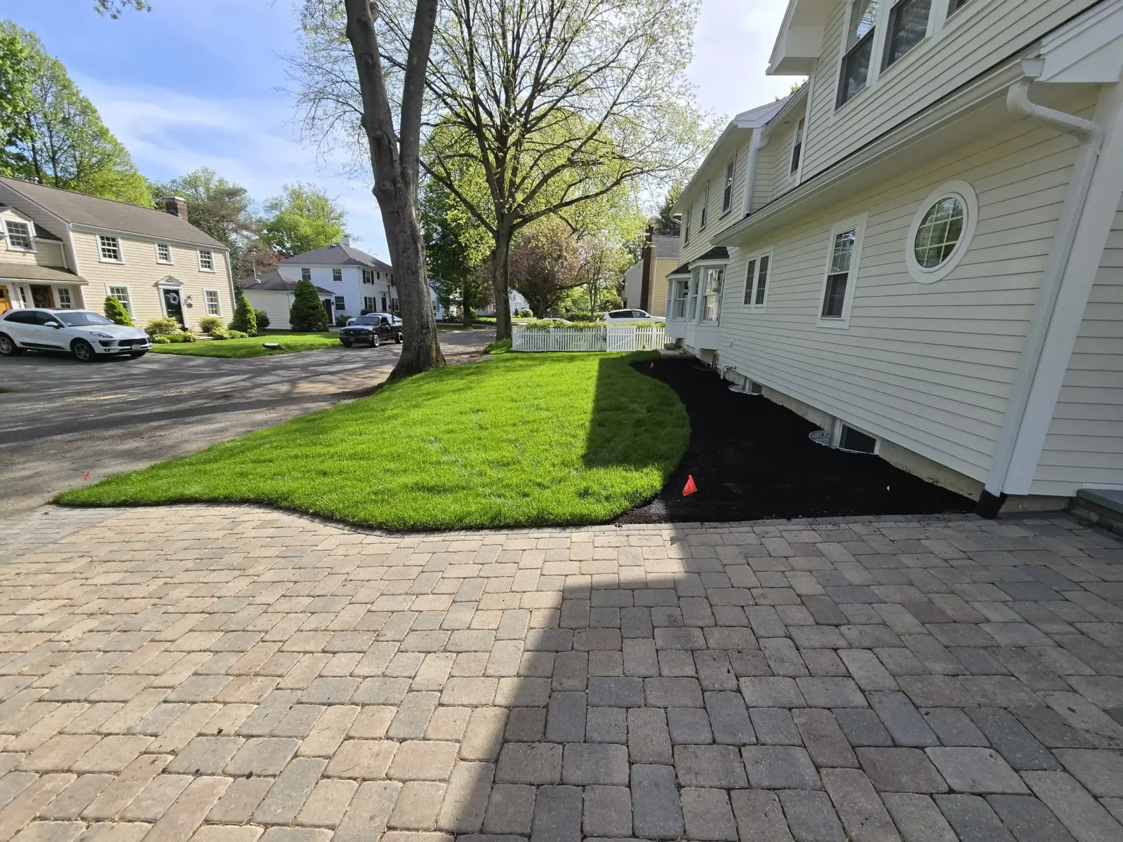 Fresh curved green lawn borders a black mulch bed beside a beige house with a brick driveway and quiet neighborhood street