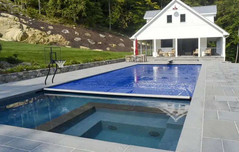 A modern backyard with a swimming pool partially covered by a blue pool cover, a built-in hot tub, a basketball hoop by the pool, and a white house with a porch in the background.