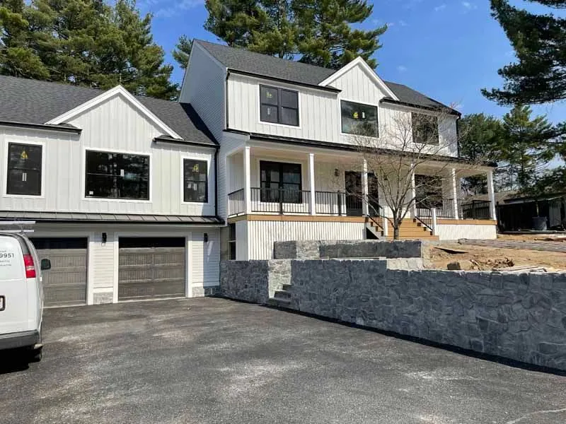 White farmhouse with black trim, gray stone retaining wall, two-car garage, and asphalt driveway.