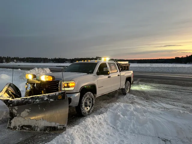 Snowplow clearing a snowy roadside at dusk, with amber lights and a pile of snow in front.