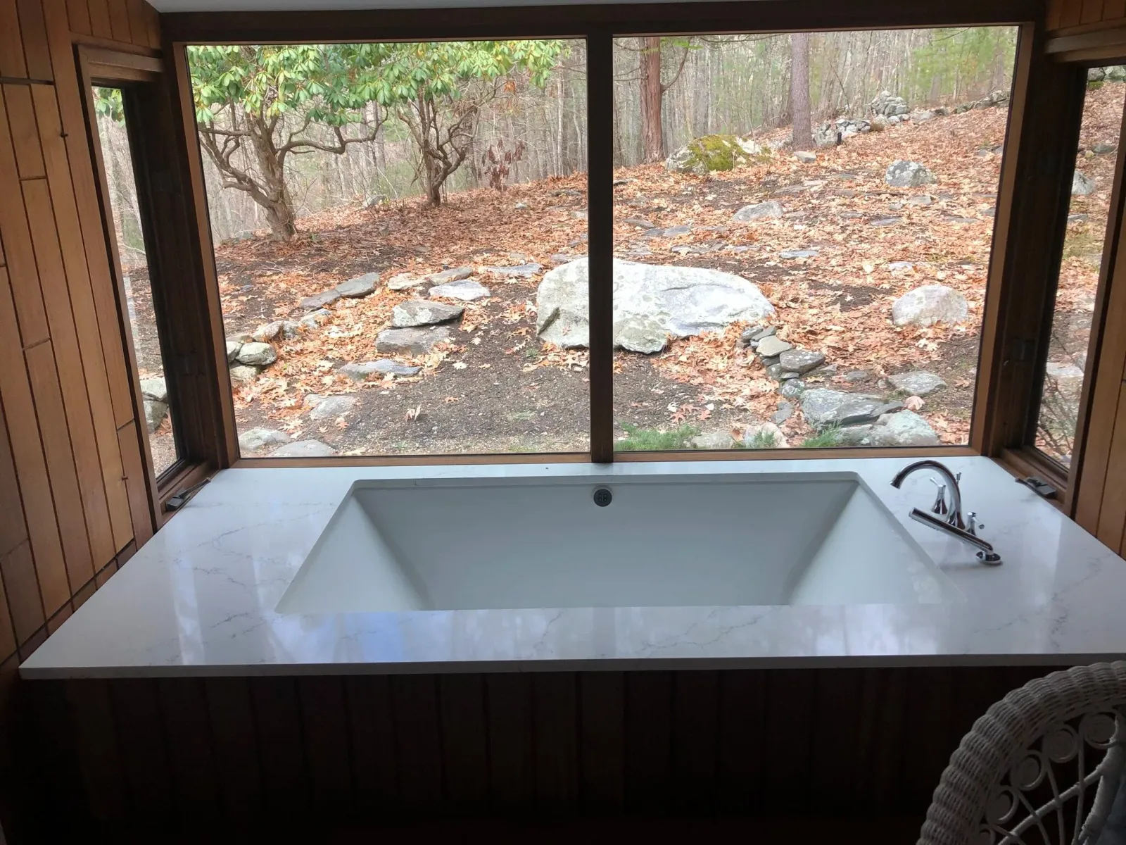 Bathtub with marble surround in a room with large windows overlooking a wooded area with rocks and trees.