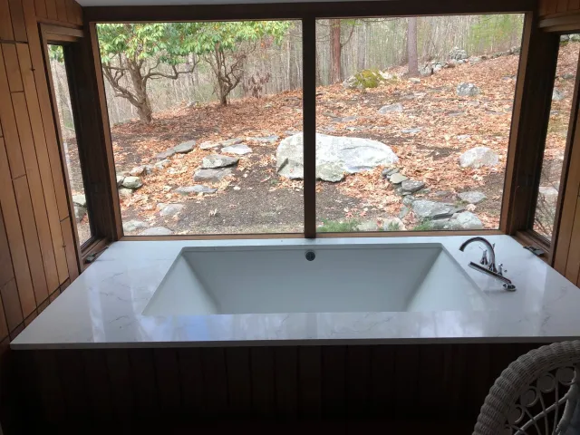 Bathtub with marble surround in a room with large windows overlooking a wooded area with rocks and trees.
