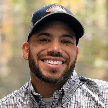 Smiling man with a beard wearing a plaid shirt and a baseball cap outdoors.