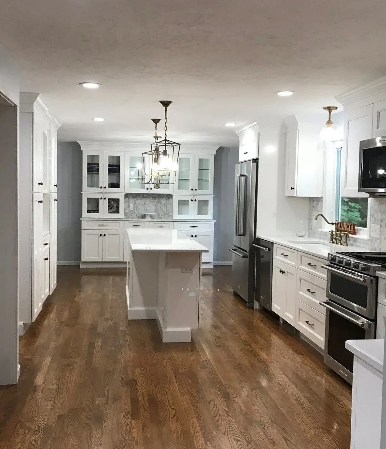 Modern white kitchen with wooden floors, stainless steel appliances, a small island, and a hanging light fixture.