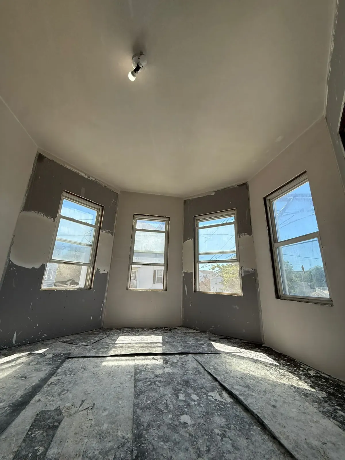 Detailed low-angle shot of a four-window bay area undergoing interior paint prep. The walls show fresh drywall compound applications and dust from sanding, while the subfloor is completely covered in a thick layer of fine sanding dust, demonstrating the scale of surface preparation in the interior paint prep workflow.