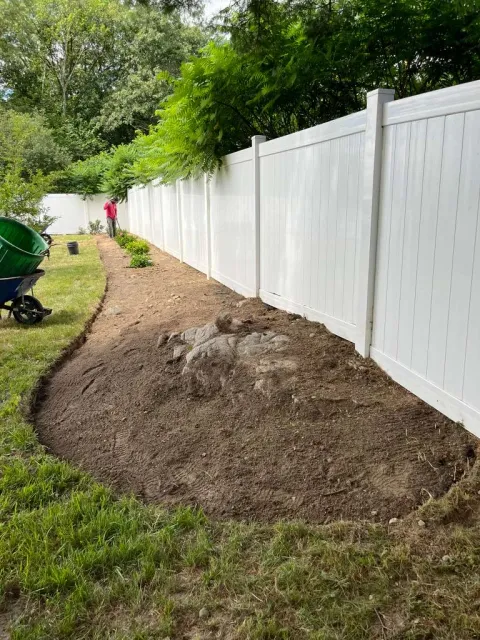 White vinyl fence along a yard with freshly turned brown soil, green trees, and a person in pink standing in the background