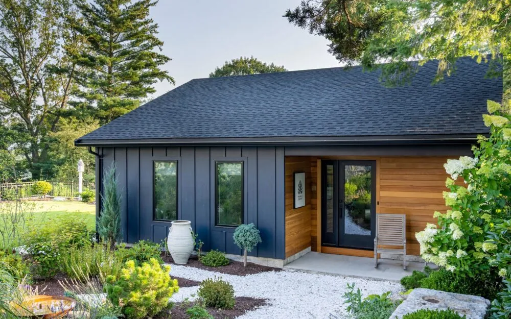 Modern house with dark blue siding, wood accents, lush greenery, and a landscaped garden – Woodland Builders, South Shore MA.