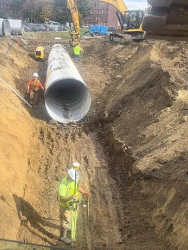Construction workers install a large corrugated drainage pipe within a deep trench at an active infrastructure project.