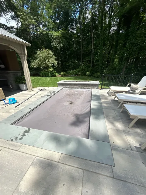 Empty hot tub covered with a beige tarp on a patio with lounge chairs and lush green trees in the background.