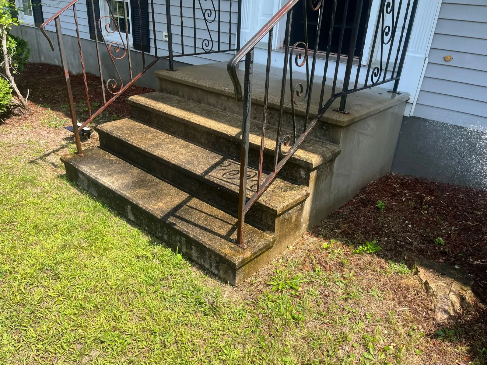 Front steps with metal railings leading to a porch beside a white house and grassy yard.