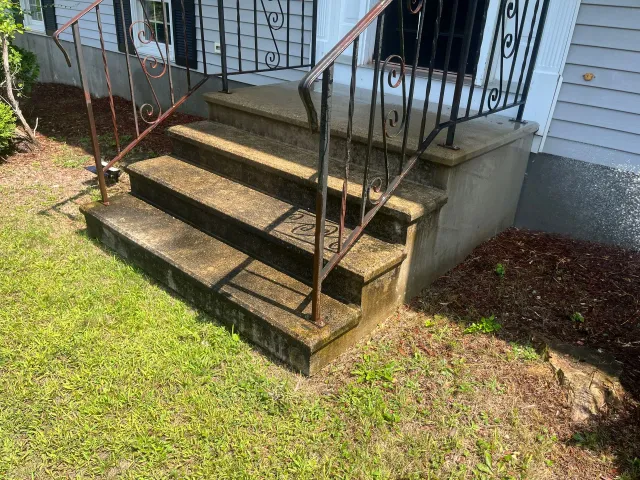 Front steps with metal railings leading to a porch beside a white house and grassy yard.
