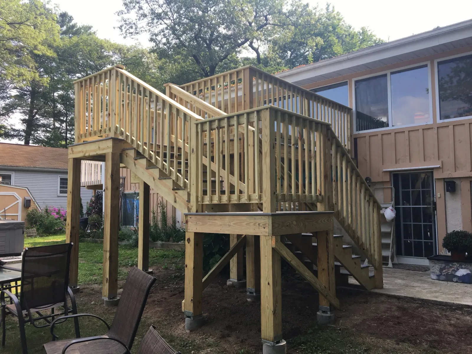 New wooden deck with stairs attached to the back of a house under construction, with outdoor chairs and a garden area in the yard.