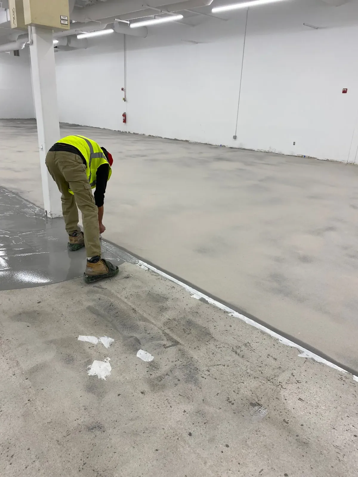 Worker in a yellow safety vest bending over a concrete floor in a large empty industrial space.