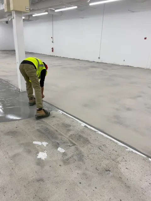 Worker in a yellow safety vest bending over a concrete floor in a large empty industrial space.